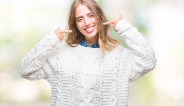 Kendinden emin gösterilen ve işaret parmakları diş ve ağız ile gülümseyen izole arka plan üzerinde kış kazak giyen güzel genç sarışın kadın. Sağlık kavramı.