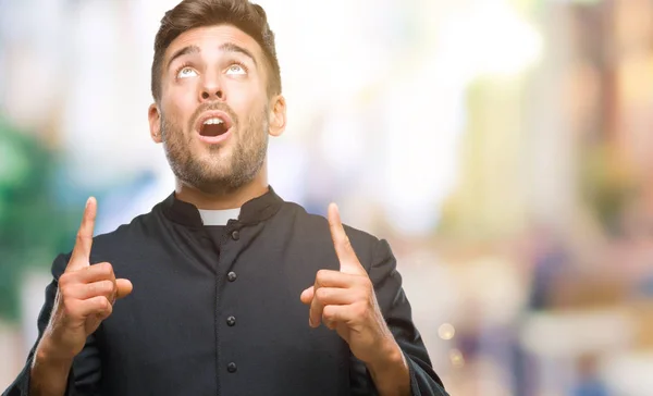 Young catholic christian priest man over isolated background amazed and surprised looking up and pointing with fingers and raised arms.