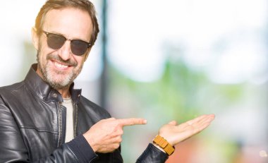 Güneş gözlüğü ve hayrete ve el ile sunulması ve parmak ile işaret ederken kameraya gülümseyen siyah deri ceket giyen orta yaş yakışıklı adam.