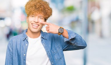 Afro saçlı kot giyen genç yakışıklı adam ceket işaretçi el parmak yüz ve burun, neşeli gülümseyen