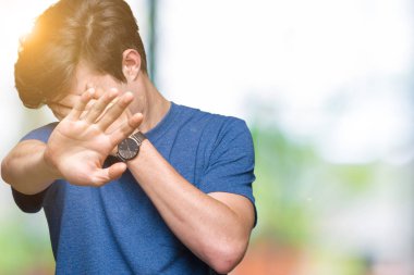 Giyen genç yakışıklı adam elleriyle gözlerini kapsayan izole arka plan üzerinde tişört mavi ve stop jest ile acı ve korku ifadesi. Utandım ve negatif kavramı.