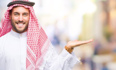 Keffiyeh neşeli sunulması ve kameraya bakarak avucunun ile işaret gülümseyen izole arka plan üzerinde giyen genç yakışıklı adam.