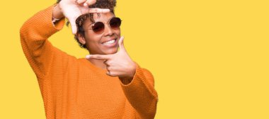 Güzel genç Afro-Amerikan kadın elle yapma çerçeve ve parmakları ile mutlu yüz gülümseyen izole arka plan üzerinde güneş gözlüğü takıyor. Yaratıcılık ve Fotoğrafçılık kavramı.