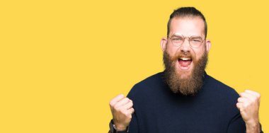 Gülümseyen ve başarı için çığlık gözlük ve balıkçı yaka kazak çok mutlu ve heyecanlı kaldırdı, silah ile kazanan jest yapıyor giyen genç sarışın adam. Kutlama kavramı.