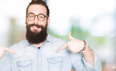 Uzun saç ve sakal yüzünde gülümseme ile kendinden emin görünüyorsun, kendini gururlu ve mutlu parmakları ile işaret gözlüklü genç hipster adamla.