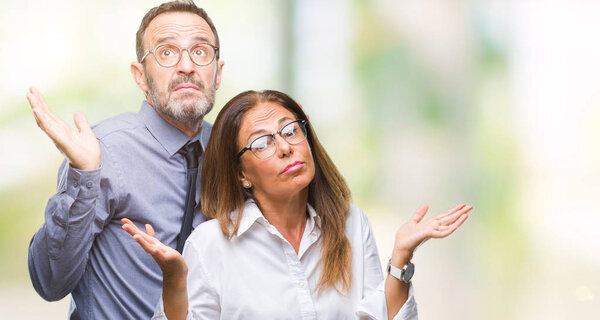 Middle age hispanic couple in love wearing glasses over isolated background clueless and confused expression with arms and hands raised. Doubt concept.