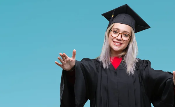 年轻的金发女子穿着毕业制服在孤立的背景看着相机张开双臂微笑拥抱 快乐的表达拥抱幸福 — 图库照片