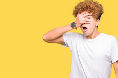 Mahcup ifadeyle parmaklarımın arasından seyir yüz ve gözler el ile kapsayan şok bakmak rahat beyaz t-shirt giyen afro saçlı Genç yakışıklı adam.