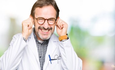 Orta yaş doktor erkekler yüksek sesle müzik gürültü rahatsız ifade parmaklarıyla kulaklarıyla kapsayan bir tıbbi palto giyiyor. Sağır kavramı.