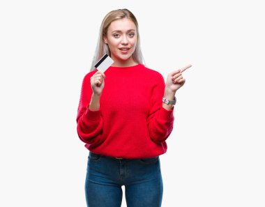 Kredi kartı izole arka plan üzerinde çok mutlu el ve parmak ile tarafına işaret tutan genç sarışın kadın