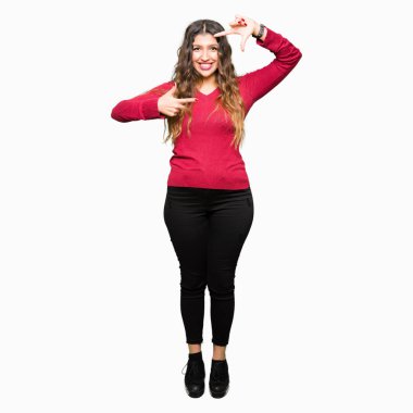 Kırmızı kazaklı elle yapma çerçeve ve parmakları ile mutlu yüz gülümseyen giyen genç güzel kadın. Yaratıcılık ve Fotoğrafçılık kavramı.