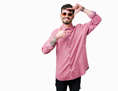 Genç yakışıklı adam elleriyle yapım çerçeve ve parmakları ile mutlu yüz gülümseyen izole arka plan üzerinde güneş gözlüğü takıyor. Yaratıcılık ve Fotoğrafçılık kavramı.