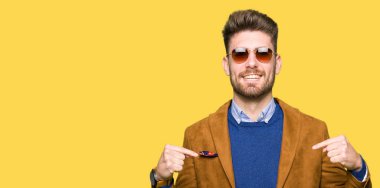 Genç yakışıklı zarif adam yüzünde gülümseme ile kendinden emin görünüyorsun, kendini gururlu ve mutlu parmakları ile işaret güneş gözlüğü takıyor.