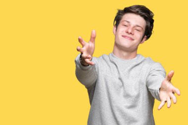 Sarılmak için kollarını açarak gülümseyerek kameraya bakarak izole arka plan üzerinde sweatshirt giyen genç yakışıklı sportif adam. Neşeli ifade kucaklayan mutluluk.