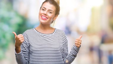 Genç güzel kadın rahat çizgili süveter izole arka plan başarı işareti el ile pozitif jest yapıyor üzerinde başparmak gülümseyen ve mutlu. Neşeli bir ifadeyle, kazanan jest kameraya bakıyor.