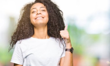 Kıvırcık saçlı hareketi el ile mutlu yaşasın yaparken rahat beyaz t-shirt giyen genç güzel kız. Başarı gösteren kameraya bakarak ifade onaylama.