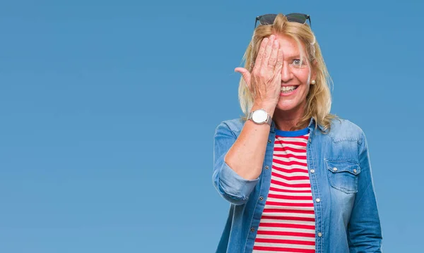 Middle age blonde woman over isolated background covering one eye with hand with confident smile on face and surprise emotion.