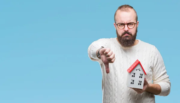 Young caucasian real state agent man holding house isolated background with angry face, negative sign showing dislike with thumbs down, rejection concept