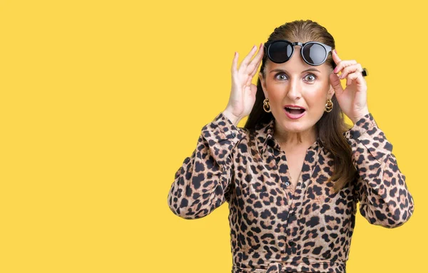 Middle age mature rich woman wearing sunglasses and leopard dress over isolated background afraid and shocked with surprise expression, fear and excited face.