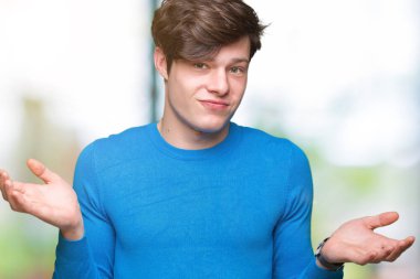 Genç yakışıklı adam izole arka plan clueless ve şaşkın bir ifadeyle kollar ve eller kaldırdı üzerinde mavi kazak giyiyor. Şüphe kavramı.