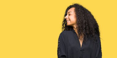 Kıvırcık saçlı uzak tarafına yüzünde, doğal ifade gülümseme ile seyir zarif elbise genç güzel kız. Kendine güvenen gülüyor.