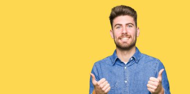 Genç yakışıklı sarışın adam el ile olumlu hareketi yaparken rahat kot gömlek başarı işareti giyiyor başparmak gülümseyen ve mutlu. Neşeli bir ifadeyle, kazanan jest kameraya bakıyor.