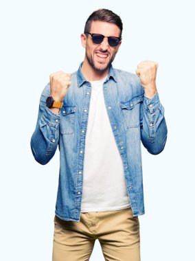 Gülümseyen ve başarı için çığlık çok mutlu ve heyecanlı kaldırdı, silah ile kazanan jest yapmak moda güneş gözlüğü takıyor yakışıklı adam. Kutlama kavramı.