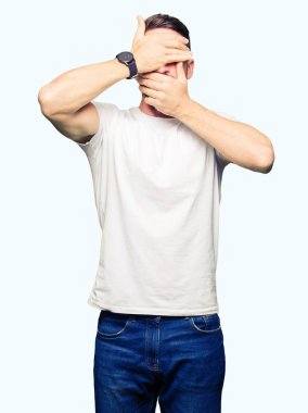 Yakışıklı adam giyiyor rahat beyaz t-shirt kapsayan gözleri ve şaşırttı ve şok ellerle ağız. Duygu gizleme