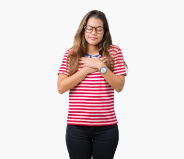 Genç güzel esmer kadın gözlük ve çizgili t-shirt üzerine arka plan ile kapalı gözler göğüs ellerinde ve yüzünde minnettar jest ile gülümseyen izole. Sağlık kavramı.
