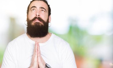 Uzun saç ve sakal yalvarıyor ve umut ifade yüzünde çok duygusal ve endişe ile birlikte elleriyle dua casual beyaz t-shirt giyen genç hipster adam. Bağışlanmak. Din kavramı.