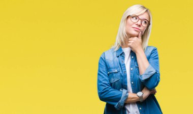 Genç güzel sarışın kadın gözlük ile izole arka plan üzerinde çene düşünme hakkında soru, dalgın ifade ver. Düşünceli yüzüyle gülümsüyor. Şüphe kavramı.