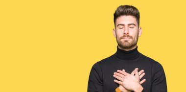 Genç yakışıklı sarışın adam ile gülümseyen kış kazak giyen kapalı gözler ve yüzünde minnettar jest ile göğüs üzerinde eller. Sağlık kavramı.