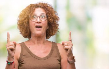 Orta yaş İspanyol kadın hayrete ve yukarıya bakıyor ve parmakları ile işaret ve silah kaldırdı sürpriz izole arka plan üzerinde gözlük.