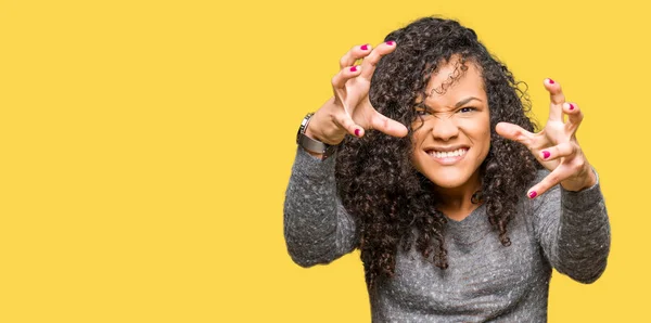 Young beautiful woman with curly hair wearing grey sweater Shouting frustrated with rage, hands trying to strangle, yelling mad