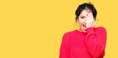 Kırmızı kazak ve el yüz ve sürpriz duygu kendinden emin gülümseme ile tek gözüyle kapsayan topuz giyen genç güzel kadın.