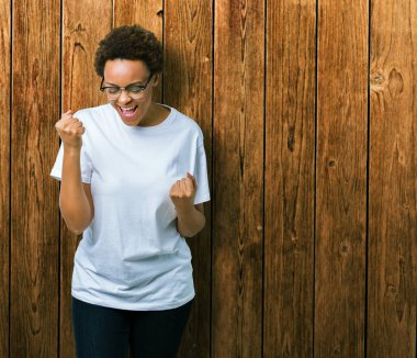 Güzel genç Afro-Amerikan kadın üzerinde gözlük gülümseyen ve başarı için çığlık arka plan çok mutlu ve heyecanlı kaldırdı, silah ile kazanan jest yapıyor izole. Kutlama kavramı.
