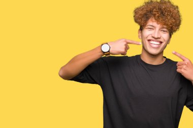 Kendinden emin gösterilen ve işaret parmakları diş ve ağız ile gülümseyen siyah t-shirt giyen afro saçlı Genç yakışıklı adam. Sağlık kavramı.