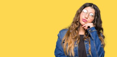 Genç güzel kadın gözlük ve denim ceket ile çene düşünme hakkında soru, dalgın ifade ver. Düşünceli yüzüyle gülümsüyor. Şüphe kavramı.