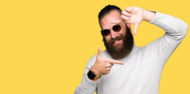 Genç hipster adam elleriyle yapım çerçeve ve parmakları ile mutlu yüz gülümseyen güneş gözlüğü takıyor. Yaratıcılık ve Fotoğrafçılık kavramı.