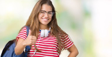 Genç güzel esmer öğrenci kadın hareketi el ile mutlu yaşasın yapıyor izole arka plan üzerinde kulaklık ve sırt çantası giyiyor. Kameraya bakarak başarı gösteren ile ifade onaylama.