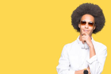 Afrikalı-Amerikalı genç afro saçlı güneş gözlüğü ve kulaklıklar el ile çene düşünme hakkında soru, dalgın ifade üzerine giyiyor. Düşünceli yüzüyle gülümsüyor. Şüphe kavramı.