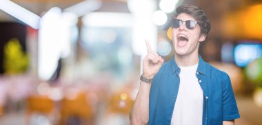 Genç yakışıklı adam başarılı fikirle izole arka plan işaret parmağı üzerinde güneş gözlüğü takıyor. Çıktıktan ve mutlu. Bir numara.