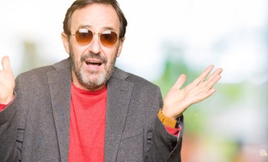Orta yaş yakışıklı iş adamı güneş gözlüğü clueless ve şaşkın bir ifadeyle kollar ve eller kaldırdı giyiyor. Şüphe kavramı.