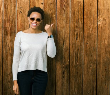 Güzel genç Afro-Amerikan kadın arıyorum ve başparmak yanında yukarıyı mutlu yüz ile gülümseyen izole arka plan üzerinde güneş gözlüğü takıyor.