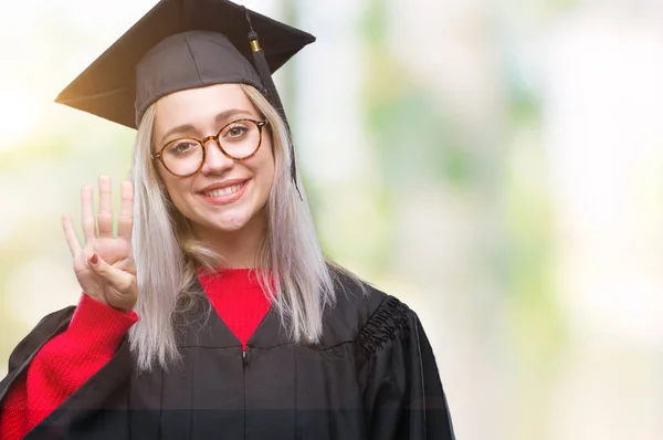 年轻的金发女子穿毕业制服在孤立的背景显示和指向与手指数字四同时微笑自信和快乐 — 图库照片