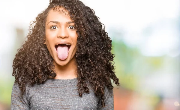 Young beautiful woman with curly hair wearing grey sweater sticking tongue out happy with funny expression. Emotion concept.