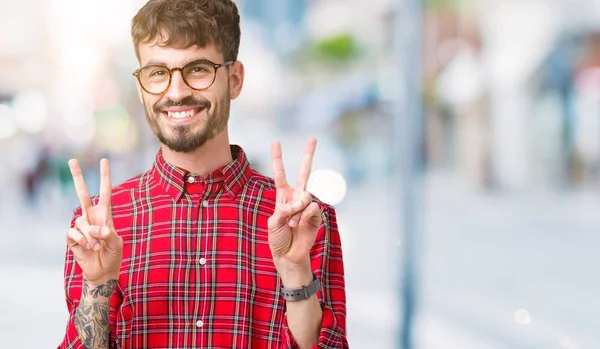 Young handsome man wearing glasses over isolated background smiling looking to the camera showing fingers doing victory sign. Number two.