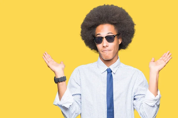 Young african american business man with afro hair wearing sunglasses clueless and confused expression with arms and hands raised. Doubt concept.