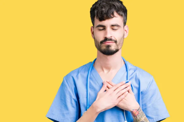 Young handsome nurse man wearing surgeon uniform over isolated background smiling with hands on chest with closed eyes and grateful gesture on face. Health concept.