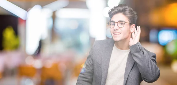 Young business man wearing glasses over isolated background smiling with hand over ear listening an hearing to rumor or gossip. Deafness concept.